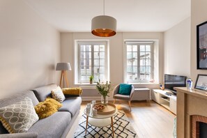 Ein modernes Wohnzimmer mit grauem Sofa, einem Couchtisch und einem Fernseher. Zwei Fenster mit weißen Rahmen bieten Blick auf umliegende Gebäude.