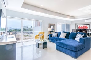 A modern living room with a glass coffee table, a blue sofa, a yellow armchair, and a large window offering a view of a construction site.