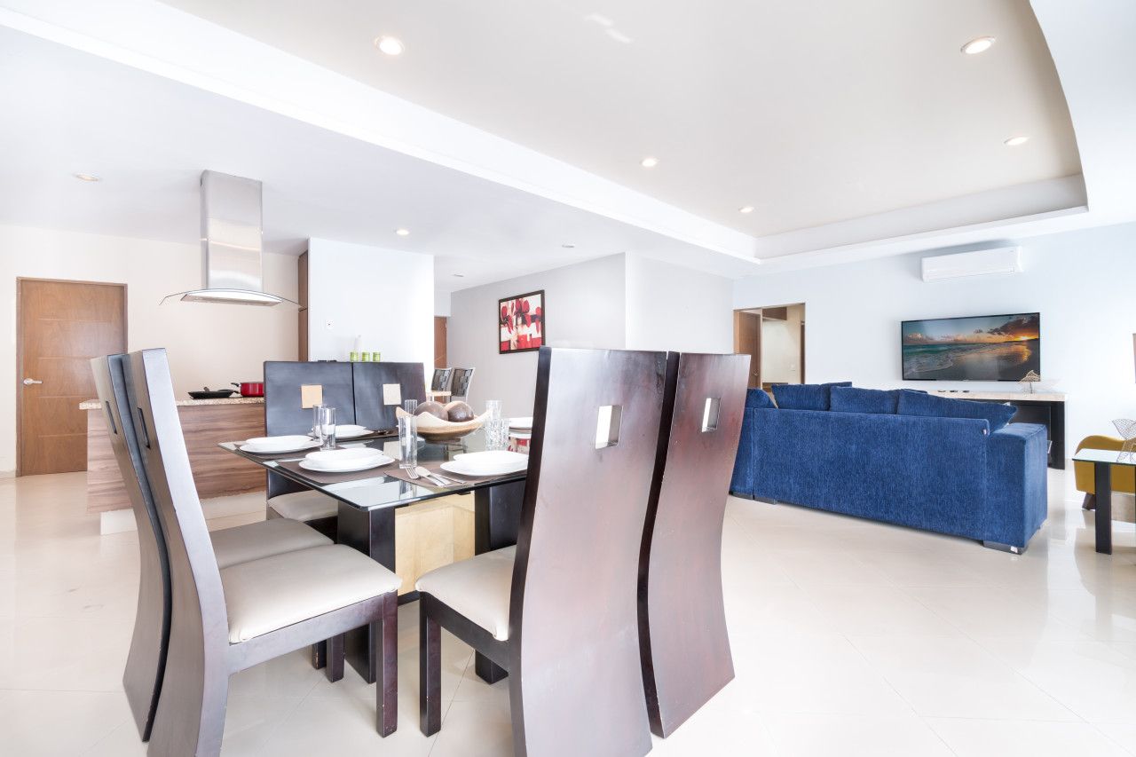A modern dining area with a kitchen in the background, a blue sofa, and a flat-screen TV mounted on the wall.