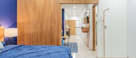 A modern bedroom with a large bed, a wooden partition, a desk with a chair, and a bathroom visible through an open door.