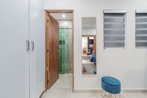 Un pasillo con una puerta de madera, ducha de vidrio, un espejo que refleja una habitación con un sofá y una alfombra, y un otoman azul.
