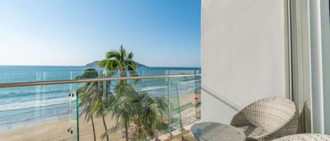 A balcony with a glass railing overlooking a beach and ocean, furnished with wicker chairs and a small table.