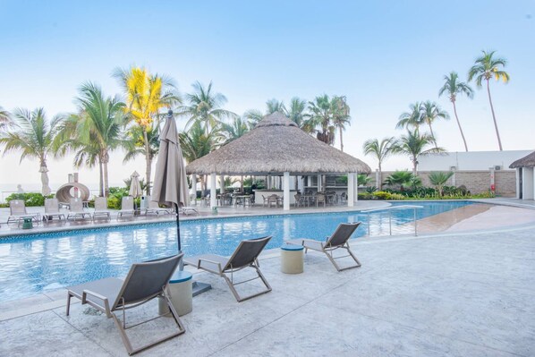 Un espace aménagé au bord de la piscine, avec des chaises longues, une structure à toit de chaume et des palmiers.