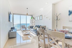 A modern living room with a sofa, dining table, and chairs, a TV mounted on the wall, and a large window offering a view of the sea.