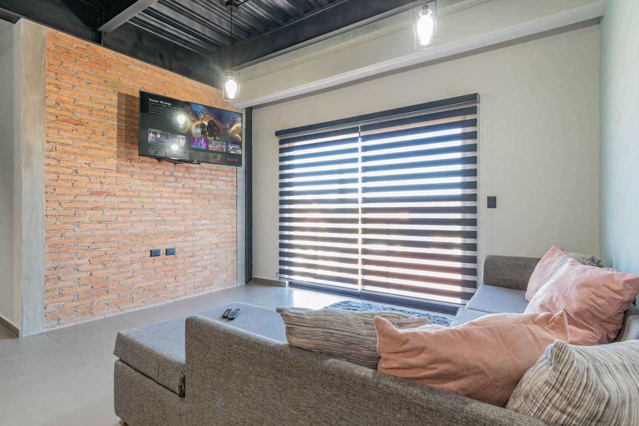 A modern living room with a brick wall, a flat-screen TV, a large window with blinds, and a grey sofa with cushions.