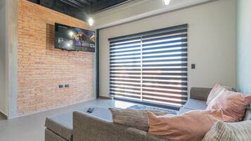 A modern living room with a brick wall, a flat-screen TV, a large window with blinds, and a grey sofa with cushions.