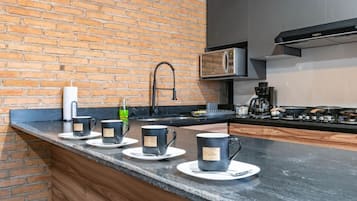 A kitchen with a brick wall, a stainless steel microwave, a coffee machine, and a black countertop.