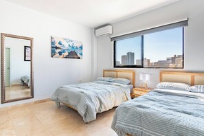 Une chambre d’hôtel avec deux lits, une grande fenêtre donnant sur la ville, un miroir et un tableau au mur.