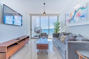 Un salon moderne avec un téléviseur à écran plat, un meuble en bois, un canapé gris, une petite table et un fauteuil suspendu près d’une grande porte vitrée donnant sur un balcon avec vue sur la mer.