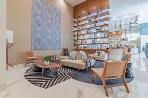 Un área de descanso moderna con un gran muro de diseño geométrico, estanterías de madera con plantas y diversas opciones de asientos, incluyendo sofás y sillones.
