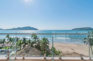 Vista da praia a partir de uma varanda, com palmeiras, uma construção de palha e céu claro.