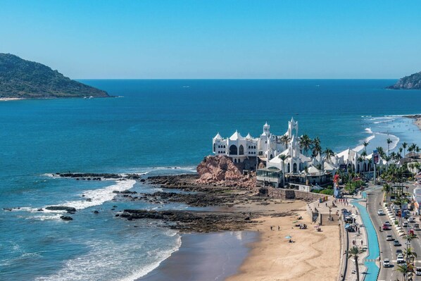 Un bâtiment situé sur la côte, doté de plusieurs tours blanches, une plage et une route où circulent des véhicules.
