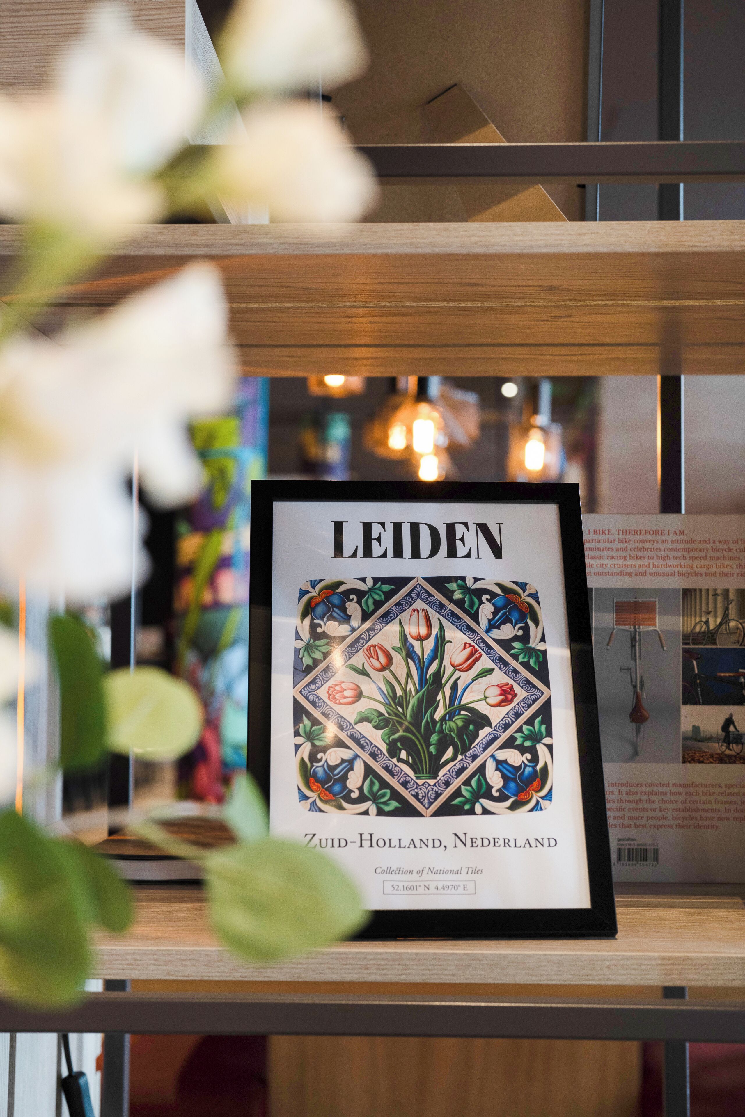 Een lijst met een tegel waarop de titel 'LEIDEN' staat, met een bloemistisch ontwerp, tentoongesteld op een plank met een onscherpe achtergrond.