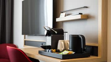 A modern hotel room with a wooden desk, a coffee machine, a tray with coffee cups and condiments, a red chair, and a television mounted on the wall.
