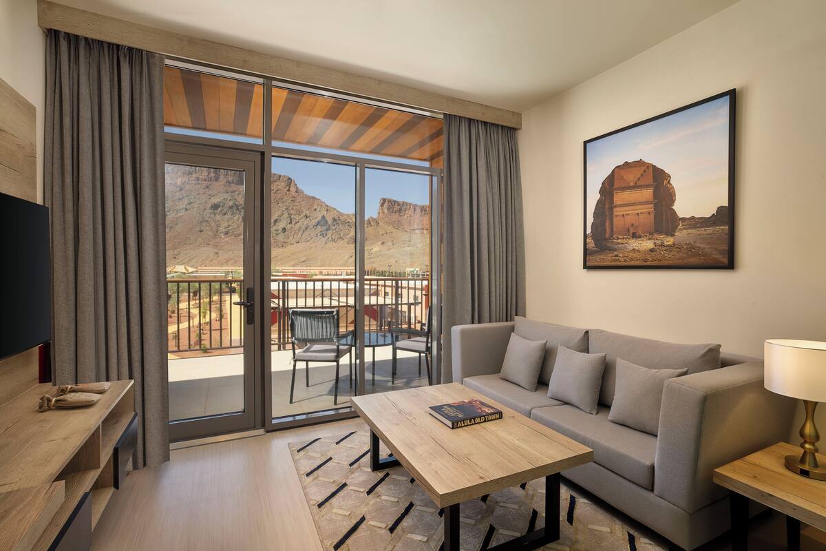 Ein modernes Wohnzimmer mit einer Couch, einem Couchtisch und einem Fernseher. Ein Balkon bietet Blick auf eine gebirgige Landschaft.
