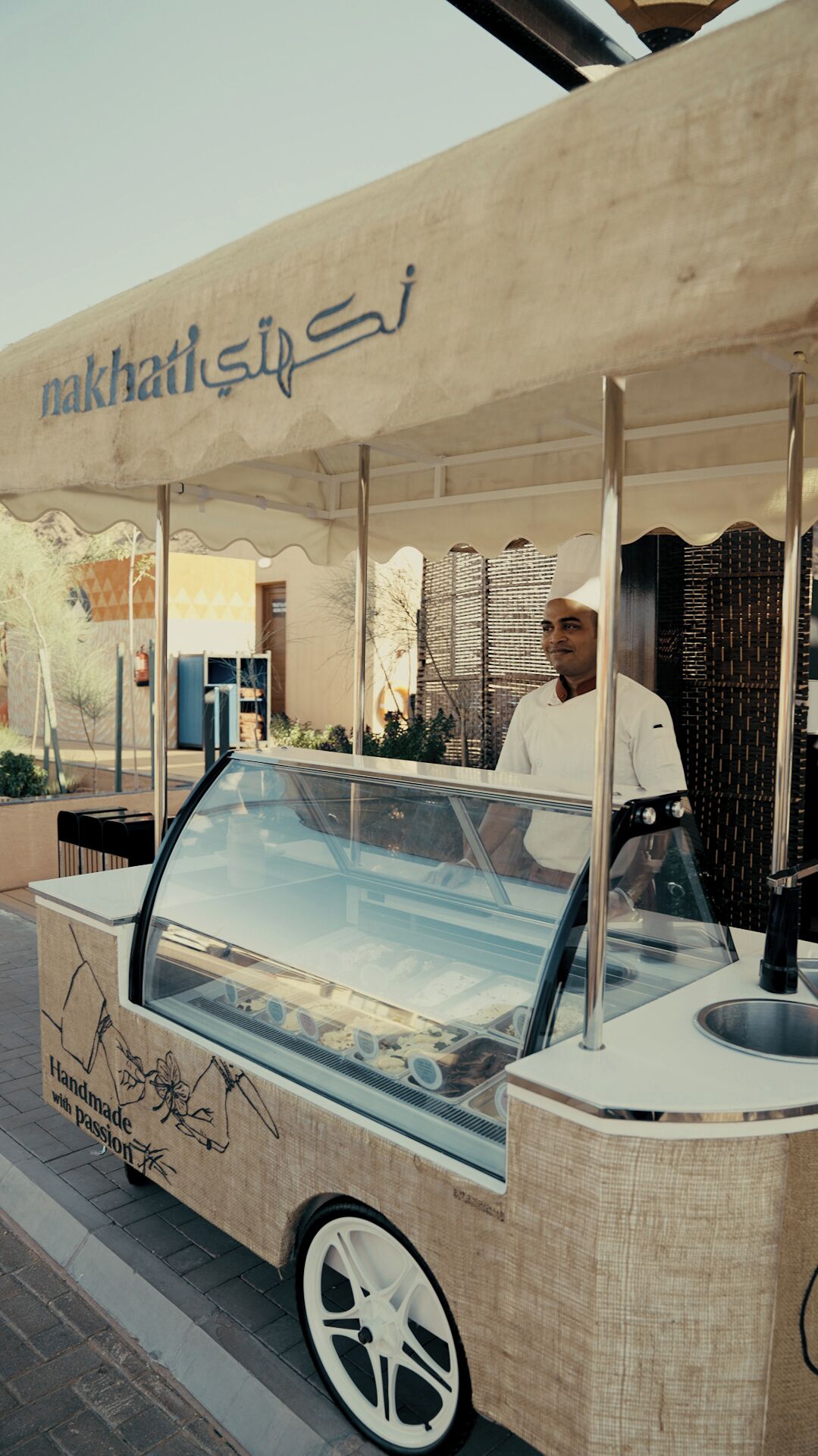 A food truck with a glass enclosure, a man standing behind it, and a sign above that reads "nakhatli".