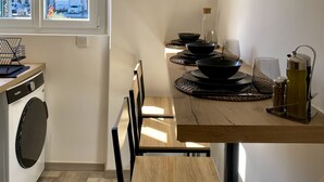 Une cuisine compacte dotée d’un lave-linge intégré, un coin repas avec une table et des chaises en bois, et une fenêtre donnant sur des immeubles.