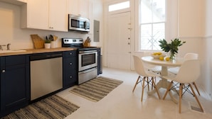 A modern kitchen with white cabinets, a microwave, oven, and dishwasher. There is a dining table with chairs and a potted plant.