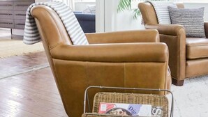 A living room with a brown leather sofa, a wicker side table with magazines, a white rug, and a large green plant.