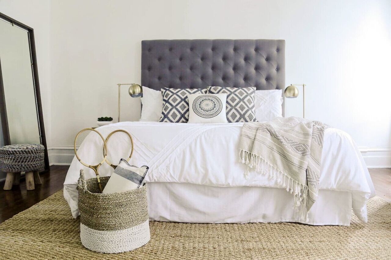 A neatly arranged bedroom with a tufted headboard, a bed with white bedding, patterned pillows, and a bedside table with a lamp.