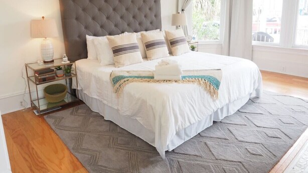 A neatly arranged bedroom with a tufted headboard, a bed with white linens and decorative pillows, a bedside table with a lamp, and a view of the outdoors through large windows. - Beautiful Forsyth Park Charmer - Amazing Location! (Savannah)