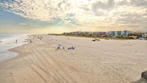 A wide sandy beach with a few people and umbrellas, buildings in the background, and a partly cloudy sky.
