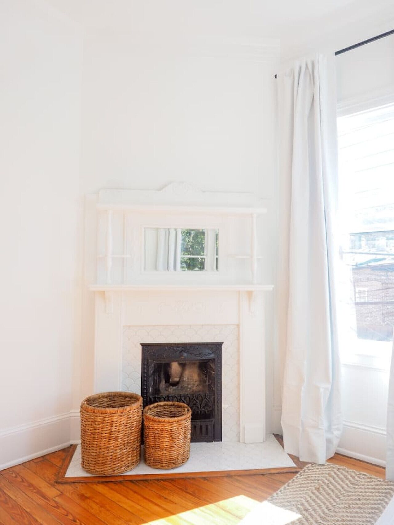A room with a white fireplace, two wicker baskets, and a view of the outside through a window with white curtains.