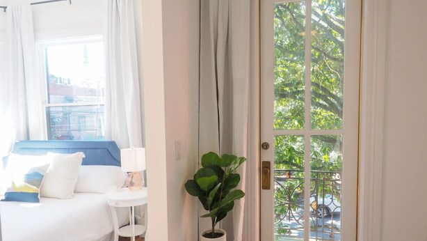 Een hotelkamer met een bed, een tafeltje, een plant in pot en een deur met uitzicht op groen buiten.