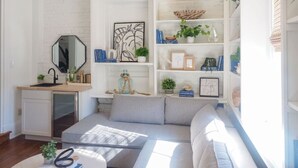 Een moderne woonkamer met een ingebouwde keuken, een grijze bank en een boekenkast vol boeken en decoratieve objecten.