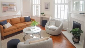 Een woonkamer met een bruine leren bank, een ronde salontafel, een grijze fauteuil en een witte schoorsteen.