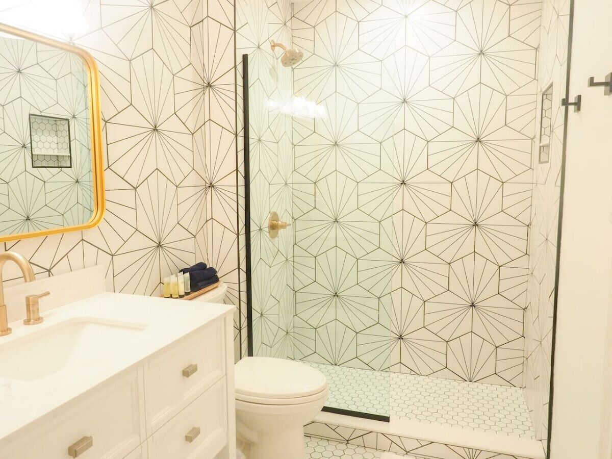 Een badkamer met een wand met een geometrisch patroon, een douchecabine van glas, een wit wastafelmeubel met gouden details en een toilet.