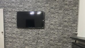 A room with a stone-patterned wall, a mounted flat-screen TV, wooden flooring, and a built-in entertainment unit with a shelf. - Tms 3bdr Tilbury - Free Parking (Tilbury)