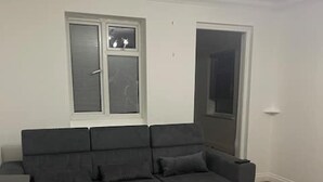 A modern living room with a dark grey sectional sofa, a wooden floor, and a window with blinds. - Tms 3bdr Tilbury - Free Parking (Tilbury)