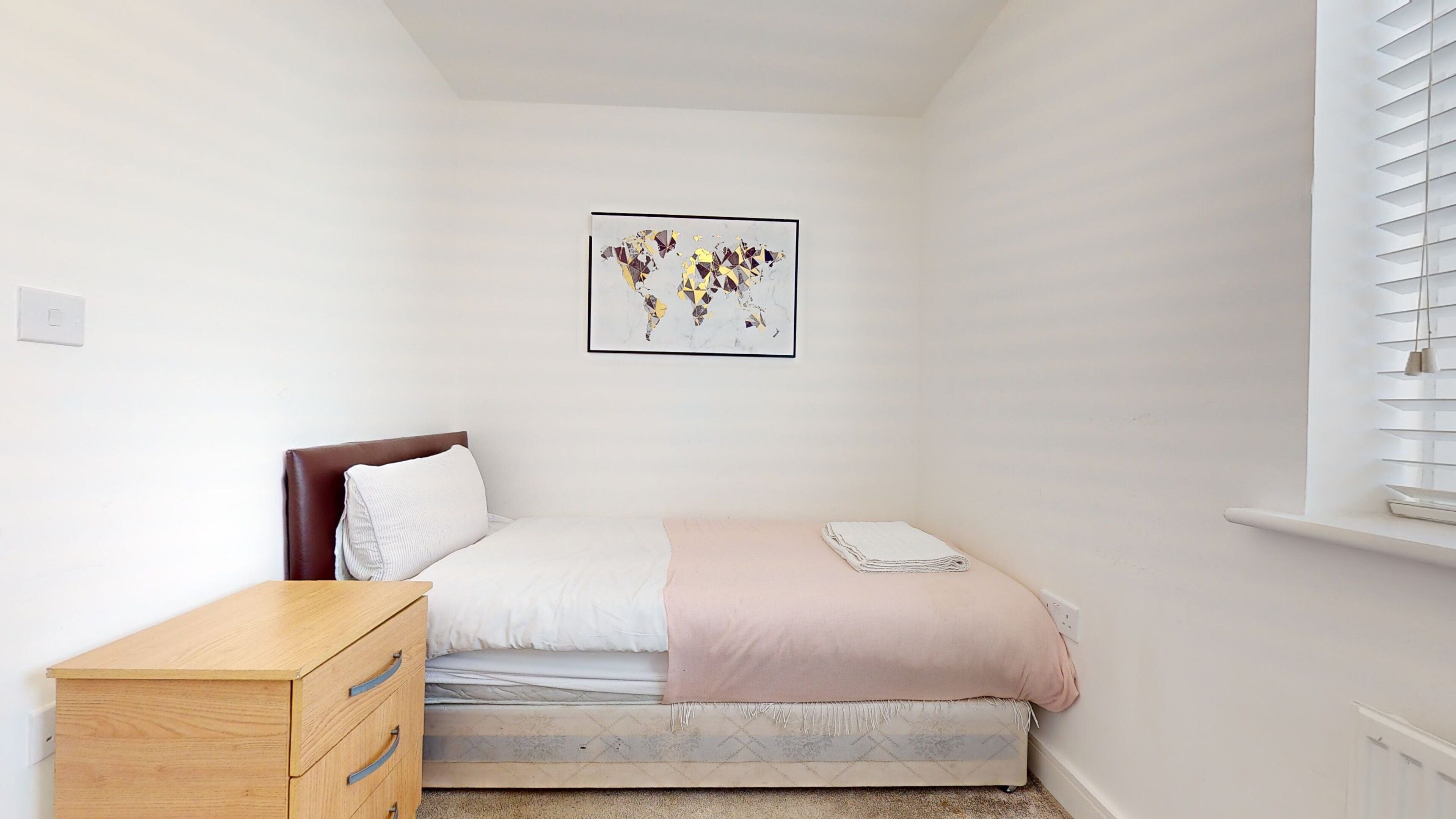 Un dormitorio con una cama, un aparador de madera, una obra de arte con un mapa mundial enmarcado y una ventana con persianas.