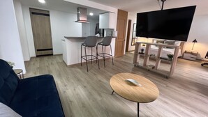 Sala de estar moderna com sofá, mesa de centro redonda de madeira e TV em um suporte. Há uma área de cozinha com micro-ondas e um banco alto.