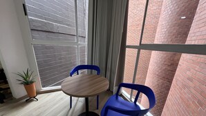 Quarto com mesa de madeira redonda, duas cadeiras azuis, uma planta em vaso e vista de um prédio de tijolos através de grandes janelas.
