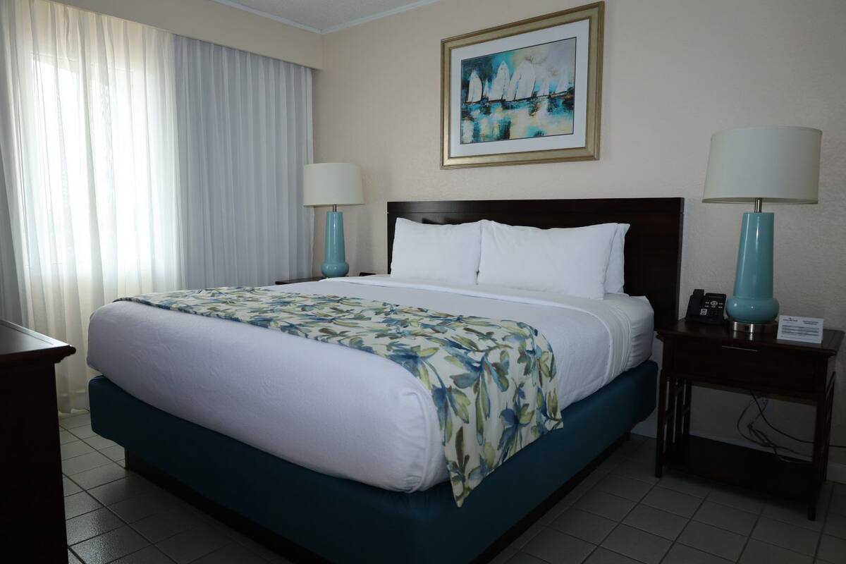 A hotel room with a large bed, two bedside lamps, a painting on the wall, and a window with curtains.