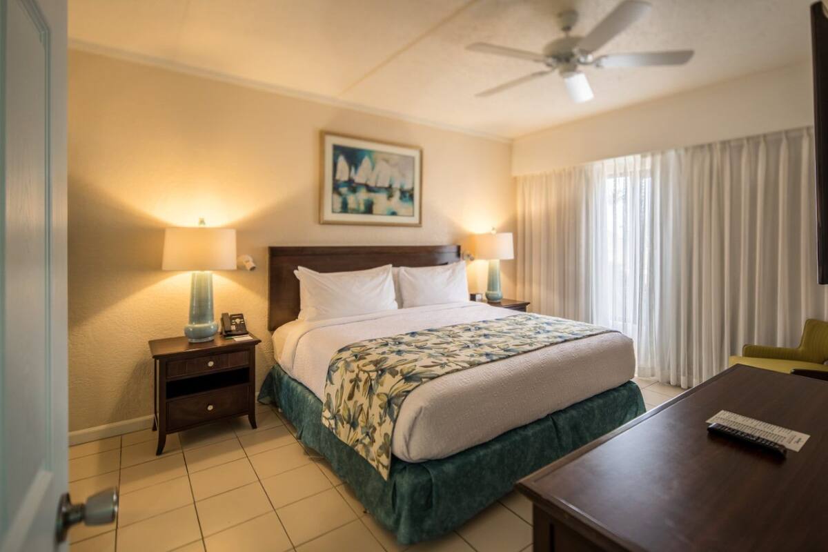 A hotel room with a bed, two bedside lamps, a ceiling fan, a painting on the wall, and a window with curtains.