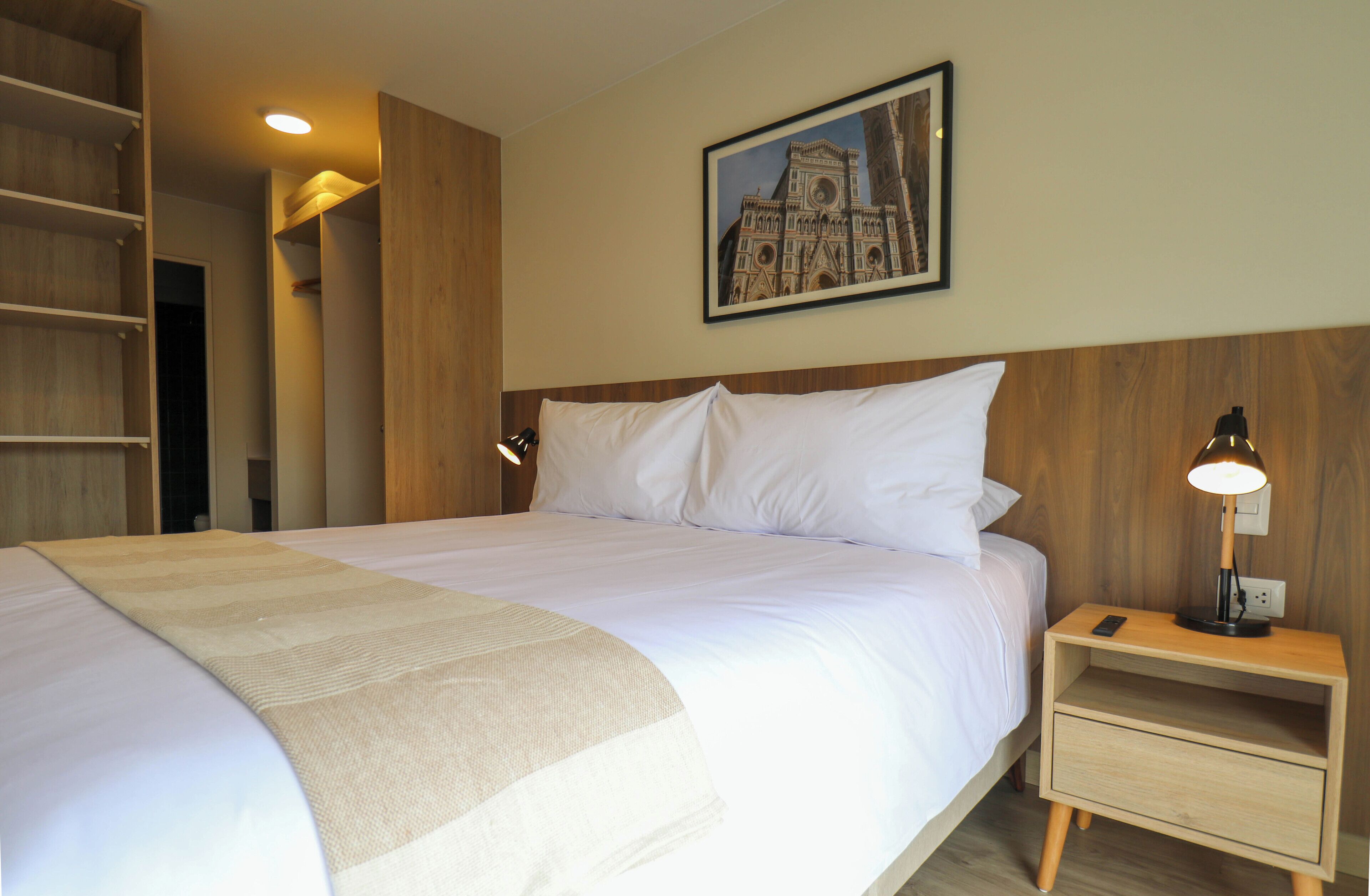 A neatly made bed with a white comforter and pillows, a bedside table with a lamp, and a framed picture of a building on the wall.