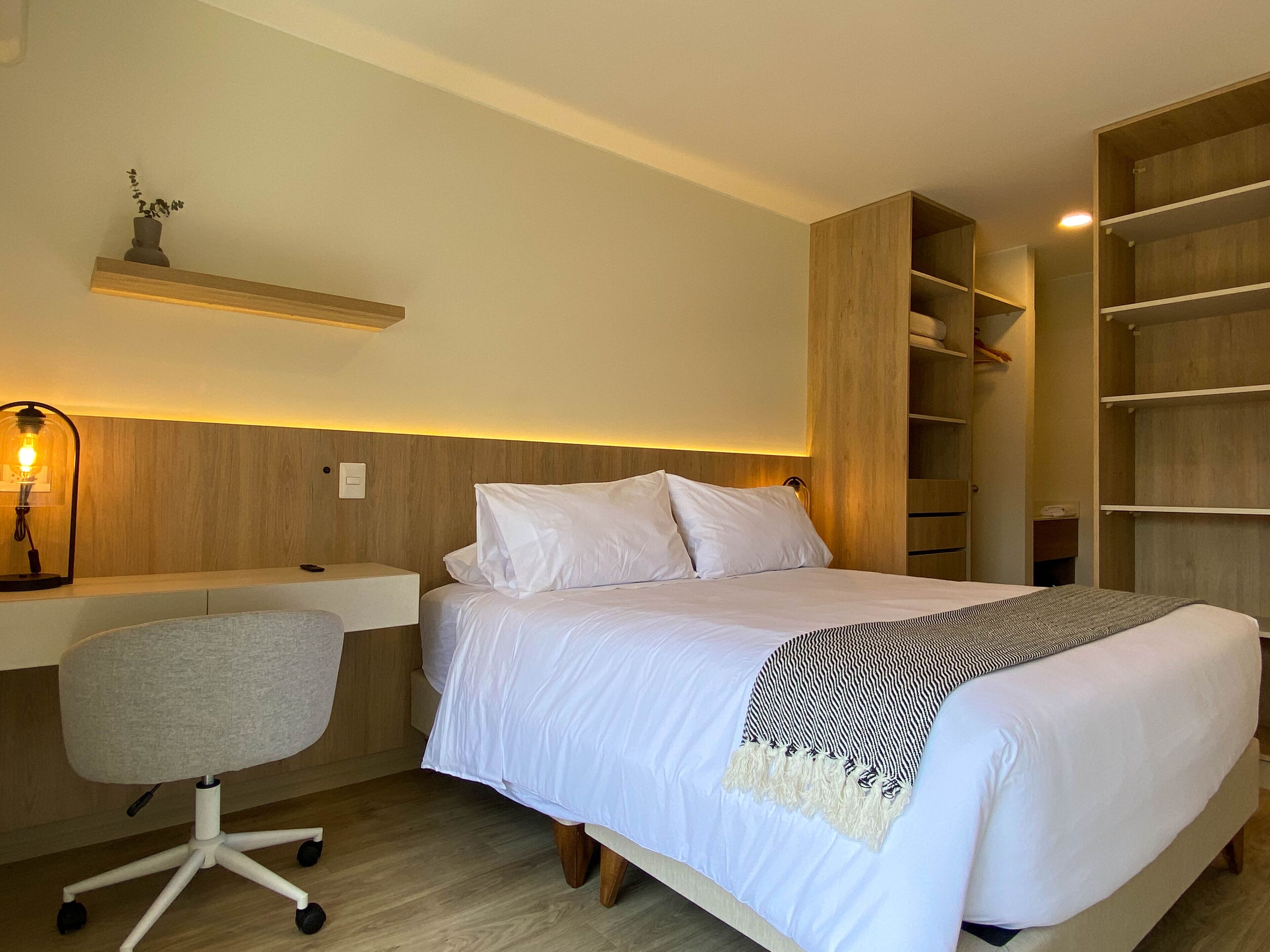 A bedroom with a bed, a desk, and a chair. There is a shelf above the bed and a wardrobe to the right.