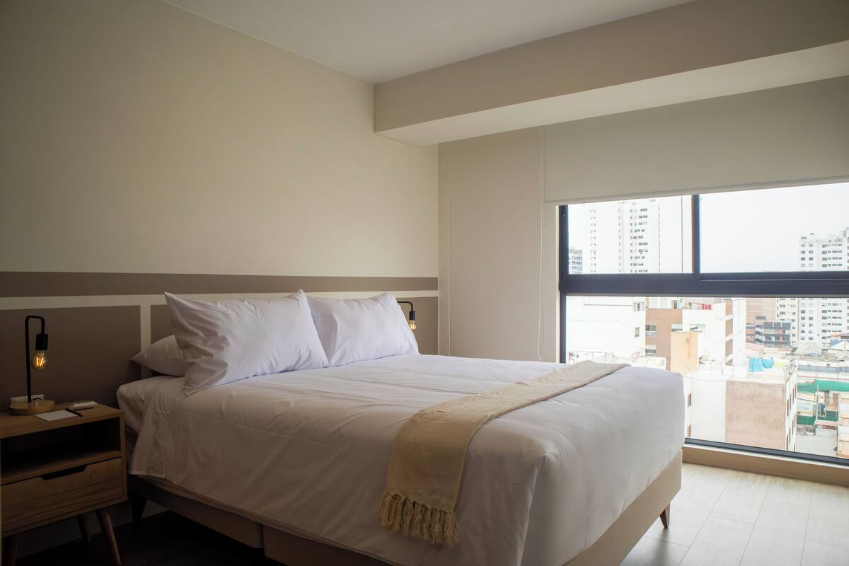 Quarto com uma cama grande, uma mesinha de cabeceira, uma luminária e janela com vista para a cidade.