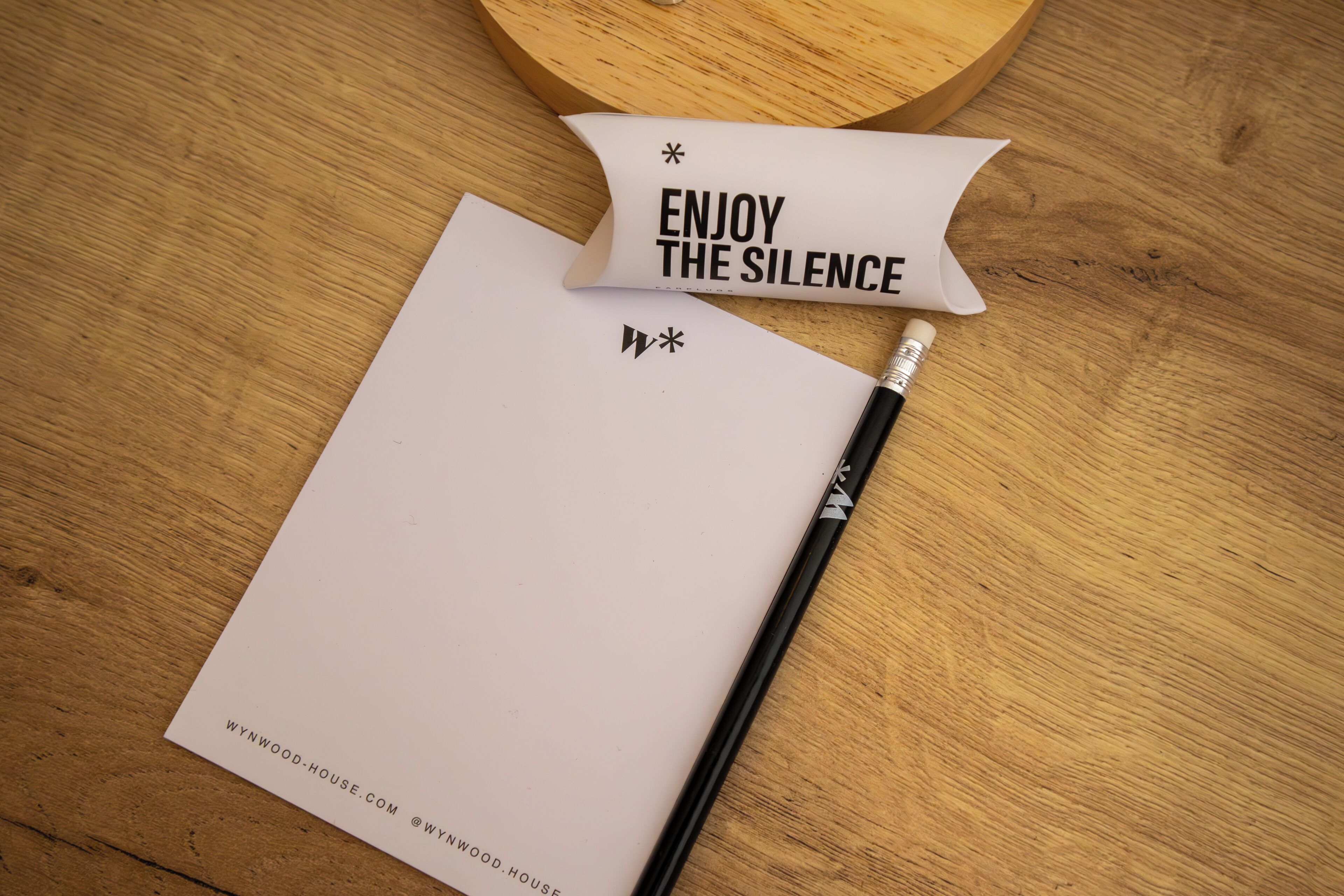 Ein Holztisch mit einem weißen Notizblock, einem schwarzen Stift und einem Kissen mit dem Aufdruck 'ENJOY THE SILENCE'.