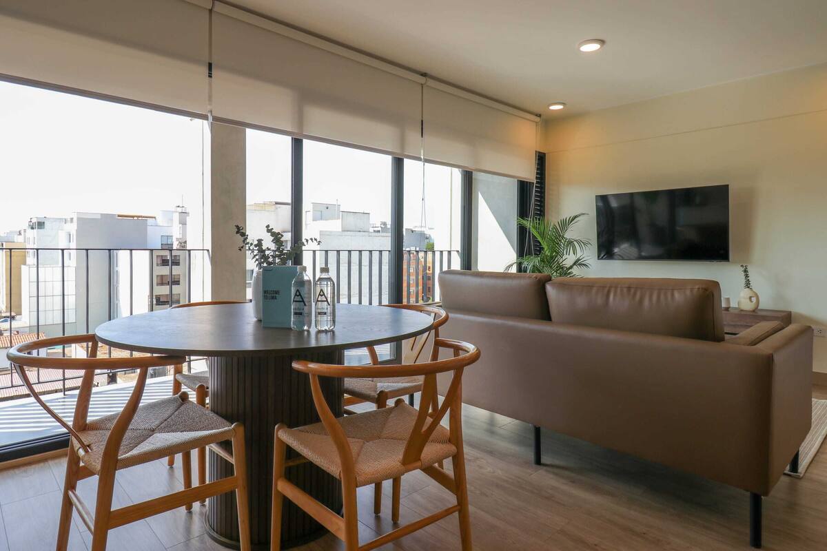 Uma sala de estar moderna com uma mesa redonda, duas cadeiras de vime, um sofá de couro marrom, uma televisão de tela plana na parede e grandes janelas com vista para a cidade.