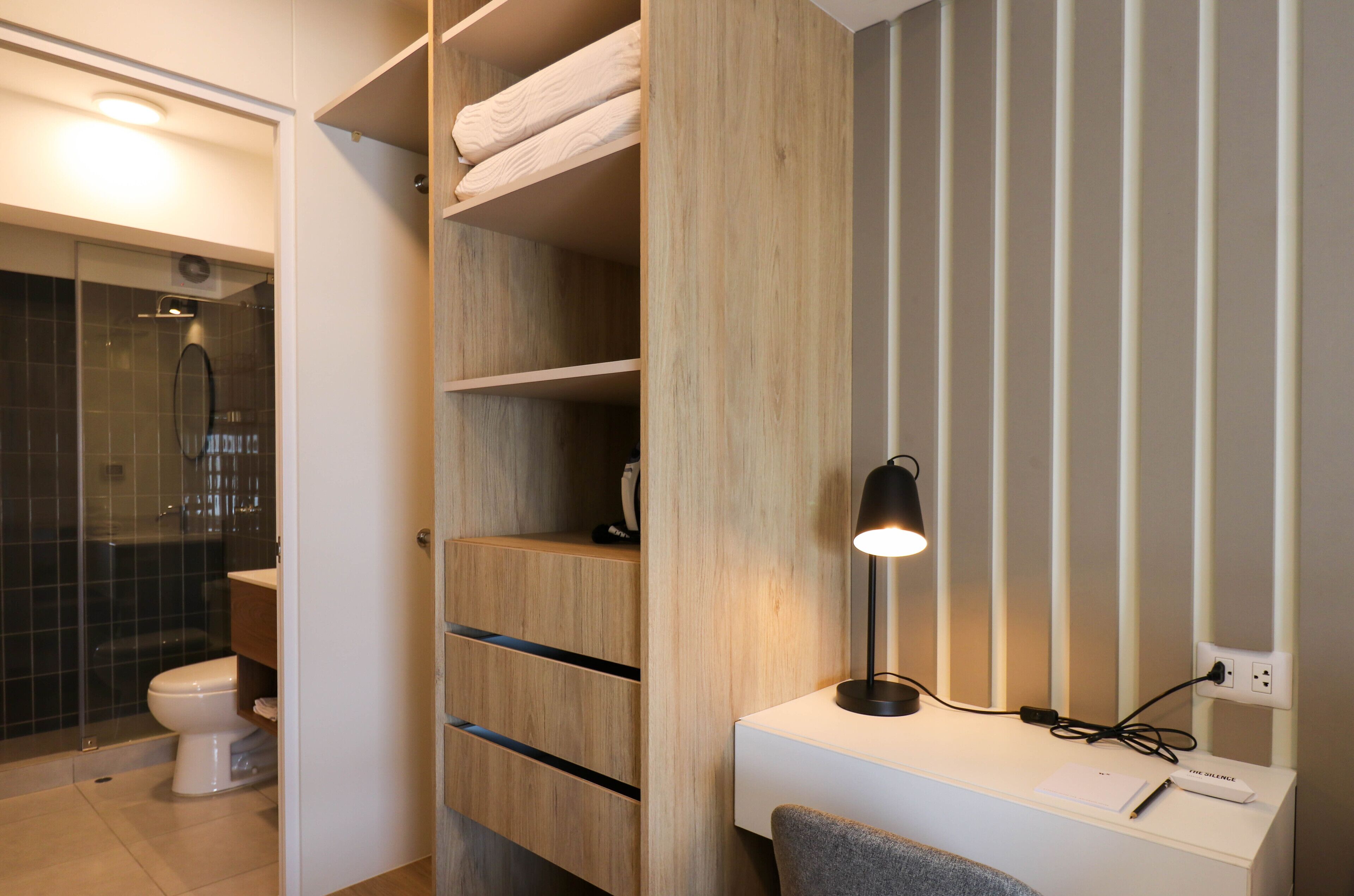 A modern bathroom with a shower, toilet, and a desk with a lamp and a notepad.