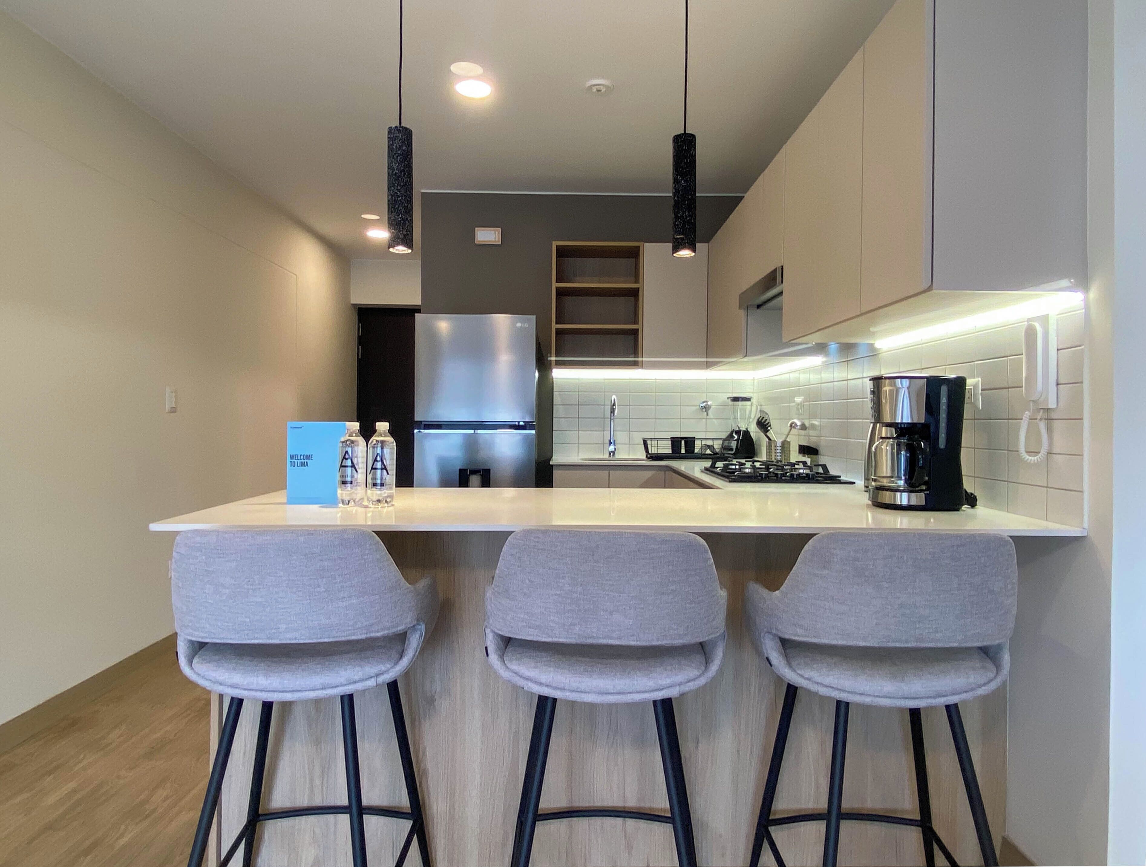 A modern kitchen with a central island, bar stools, a refrigerator, and a coffee maker.