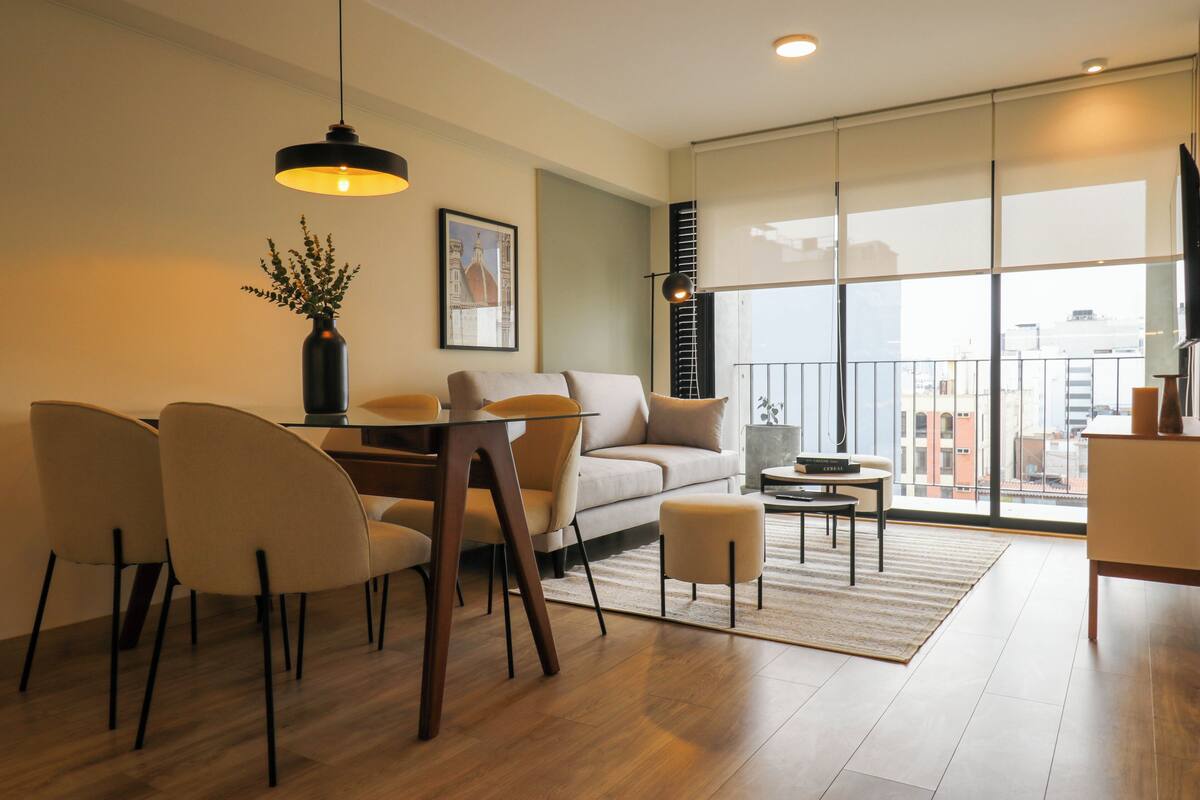 Uma sala de estar moderna com sofá, mesa de jantar, cadeiras e uma janela grande com vista para edifícios.