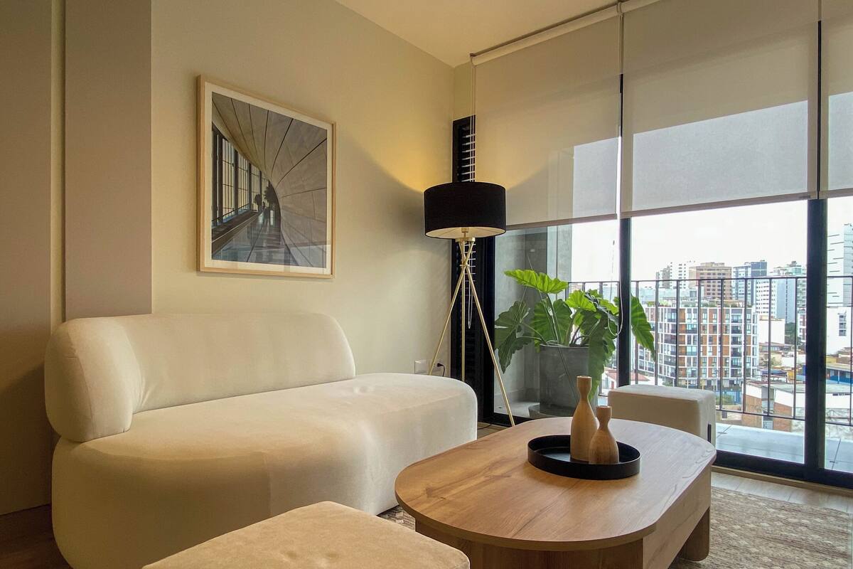 Uma sala de estar moderna com um sofá branco, uma mesa de centro de madeira e uma janela grande com vista para a cidade.