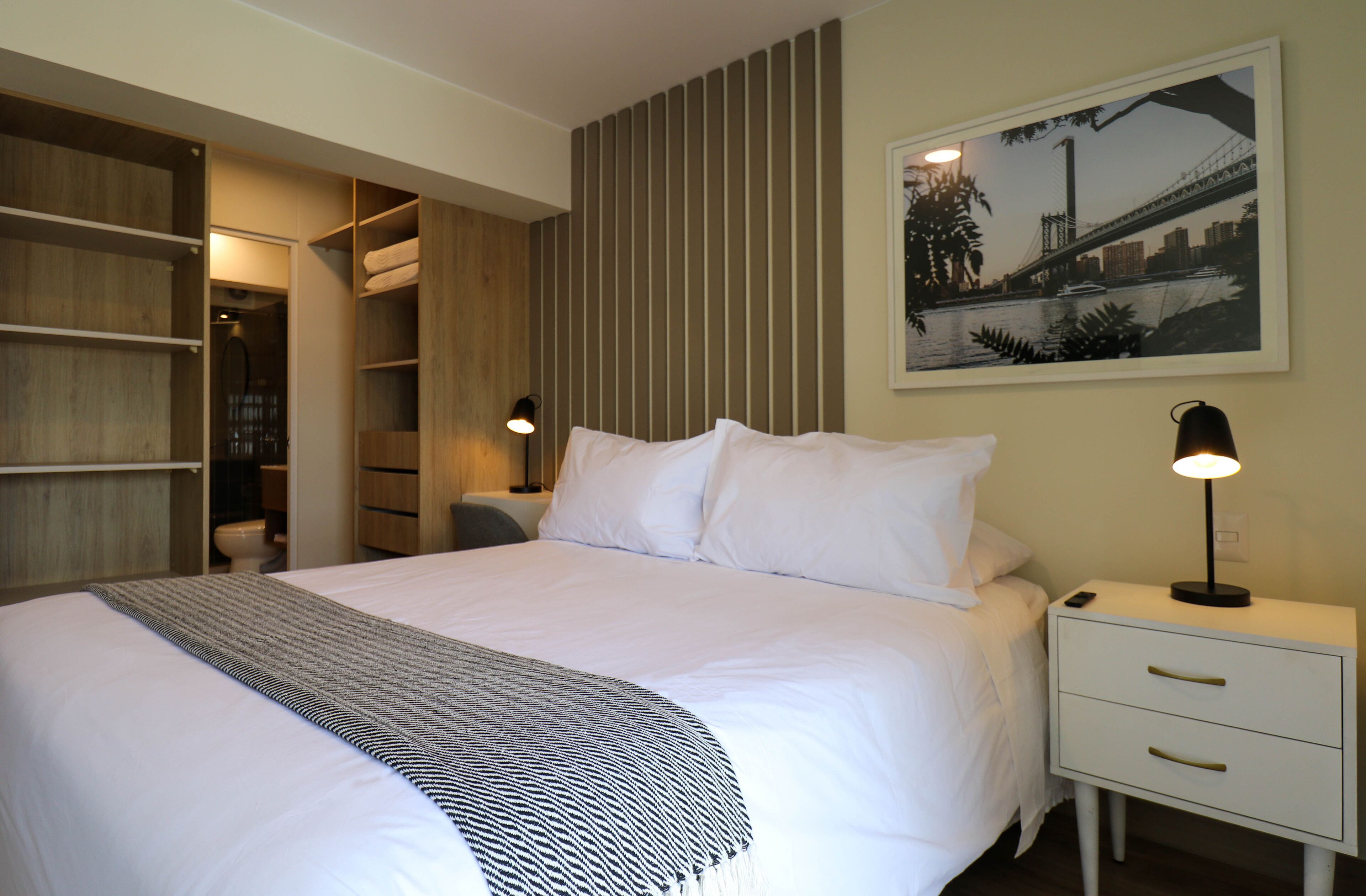 A neatly made bed with a patterned throw, a bedside table with a lamp, and a framed picture of a bridge on the wall.