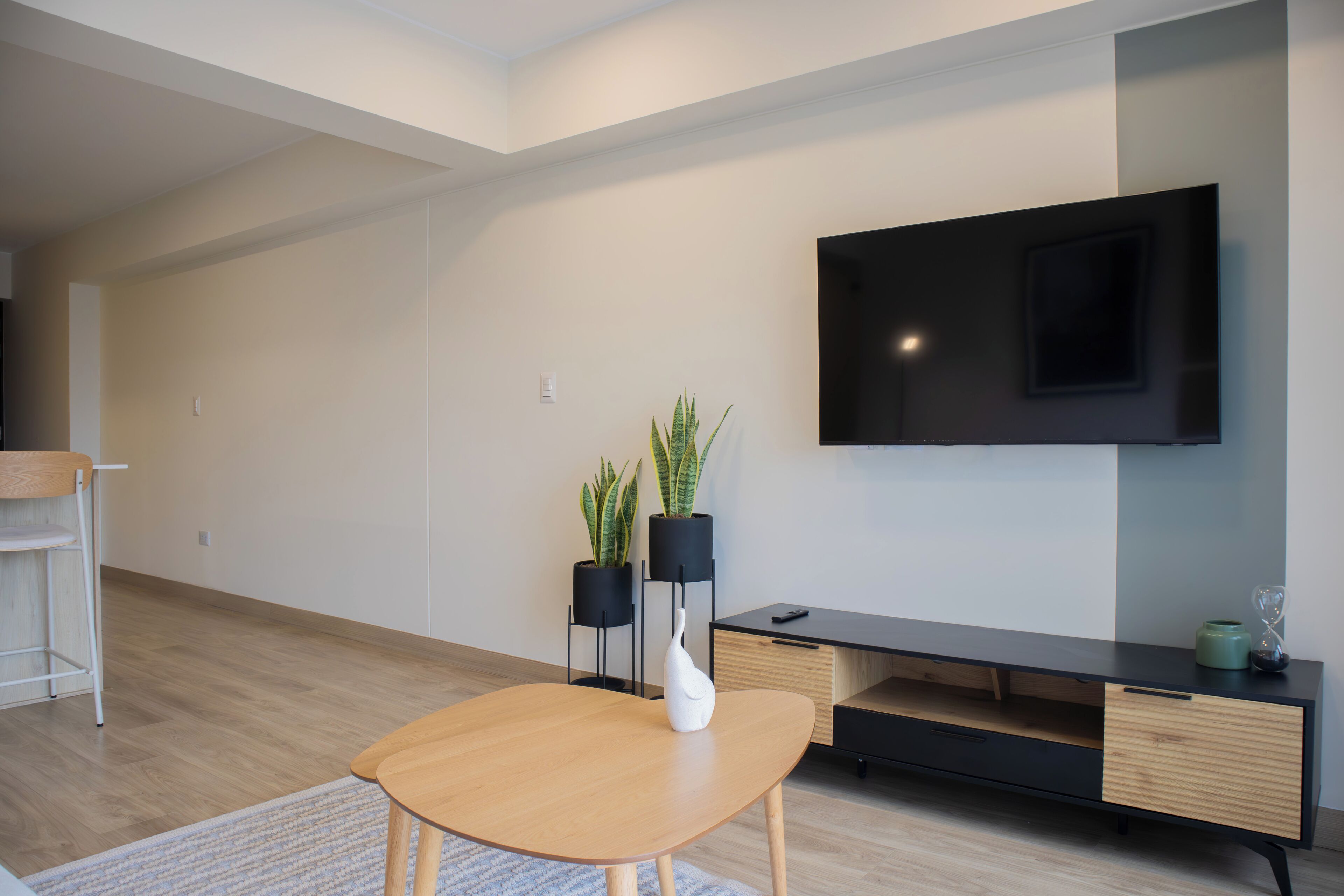 A modern living room with a flat-screen TV mounted on the wall, a wooden dining table with a white vase, and two potted plants.
