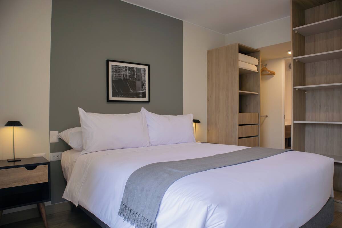 Uma cama bem arrumada com um cobertor cinza, uma mesa de cabeceira com abajur, um quadro na parede e um guarda-roupa de madeira com espaço para pendurar roupas.